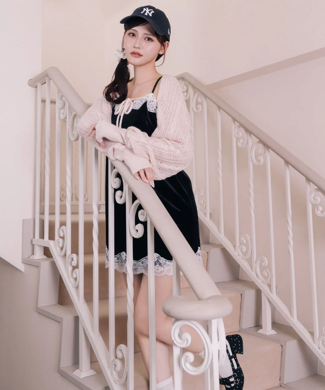 黒いベロアのキャミワンピースとピンクのニットボレロを着た女性が、NYロゴのキャップを被り、白い手すりの階段に立っています。