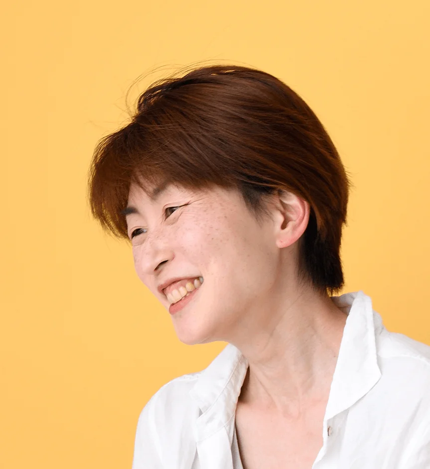 黄色い背景の前で、横向きに明るく笑うショートヘアの女性のポートレート