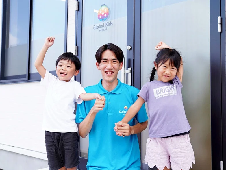 笑顔の男性教師と2人の子供たちが、Global Kids methodの建物の前で楽しそうにポーズ