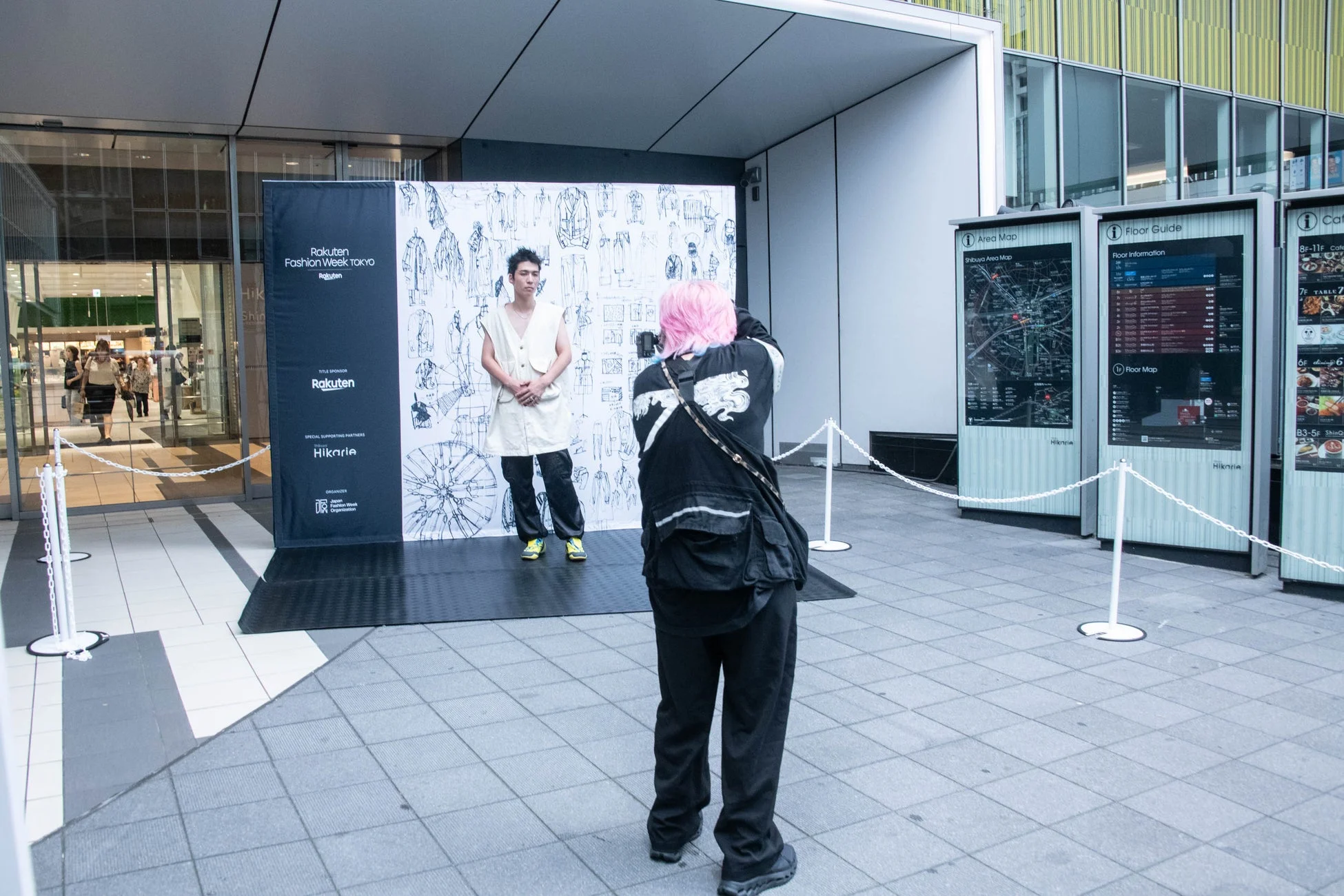 楽天ファッションウィーク東京のイベント会場で、モデルがバナーの前でポーズを取り、ピンク髪のフォトグラファーが撮影している様子