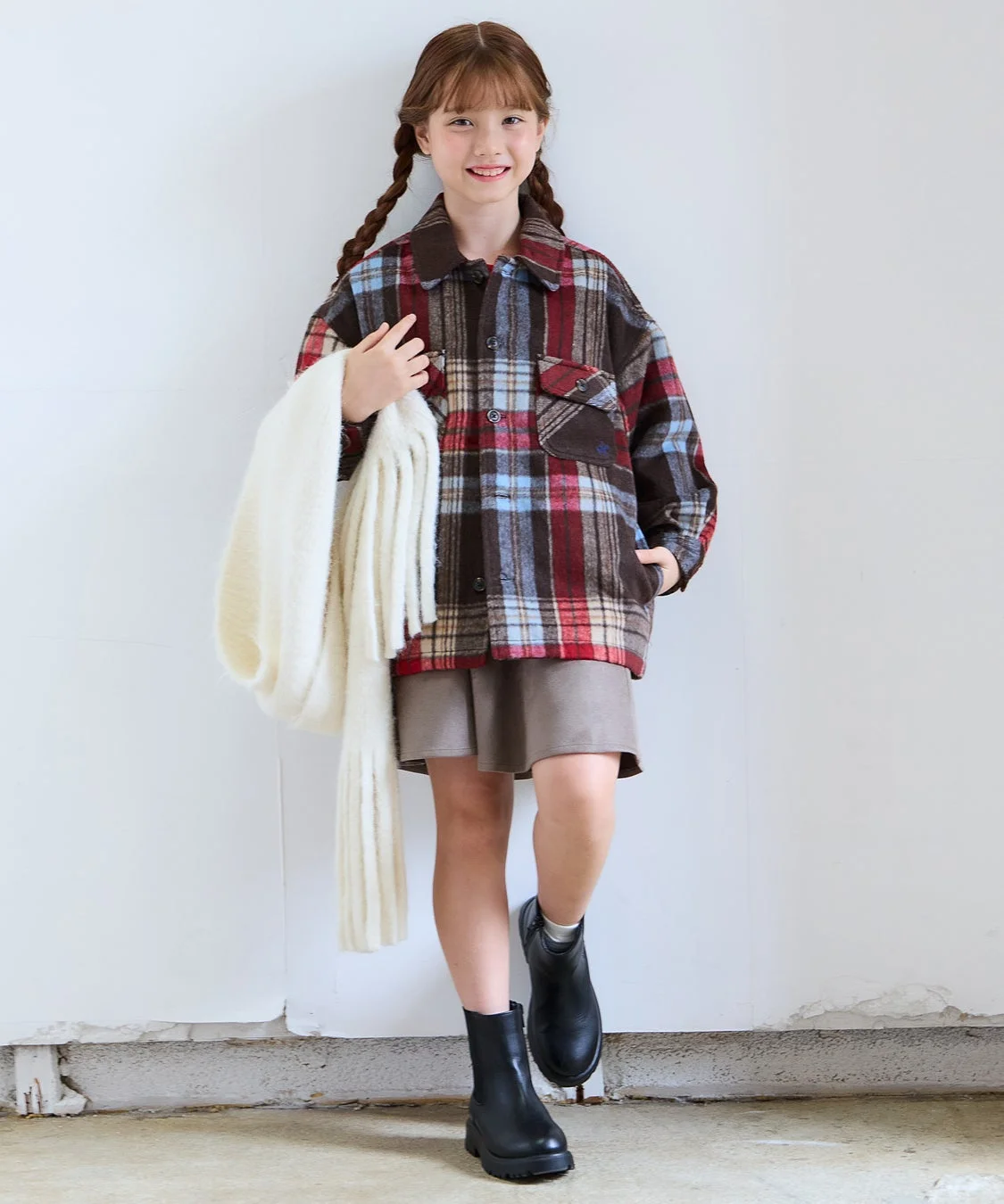 おさげ髪の女の子が、チェック柄のシャツとショートパンツにブーツを合わせたカジュアルな装いで立っている