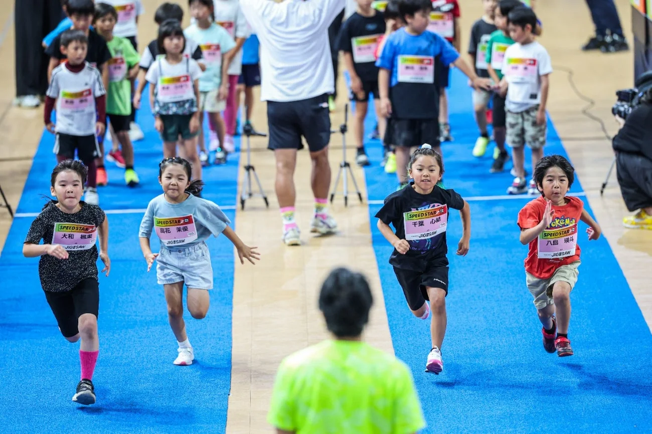 子供たちが室内トラックで徒競走を楽しんでいる様子。ゼッケンをつけた数人の子供たちが懸命に走り、背景には他の参加者や大人たちが見えます。