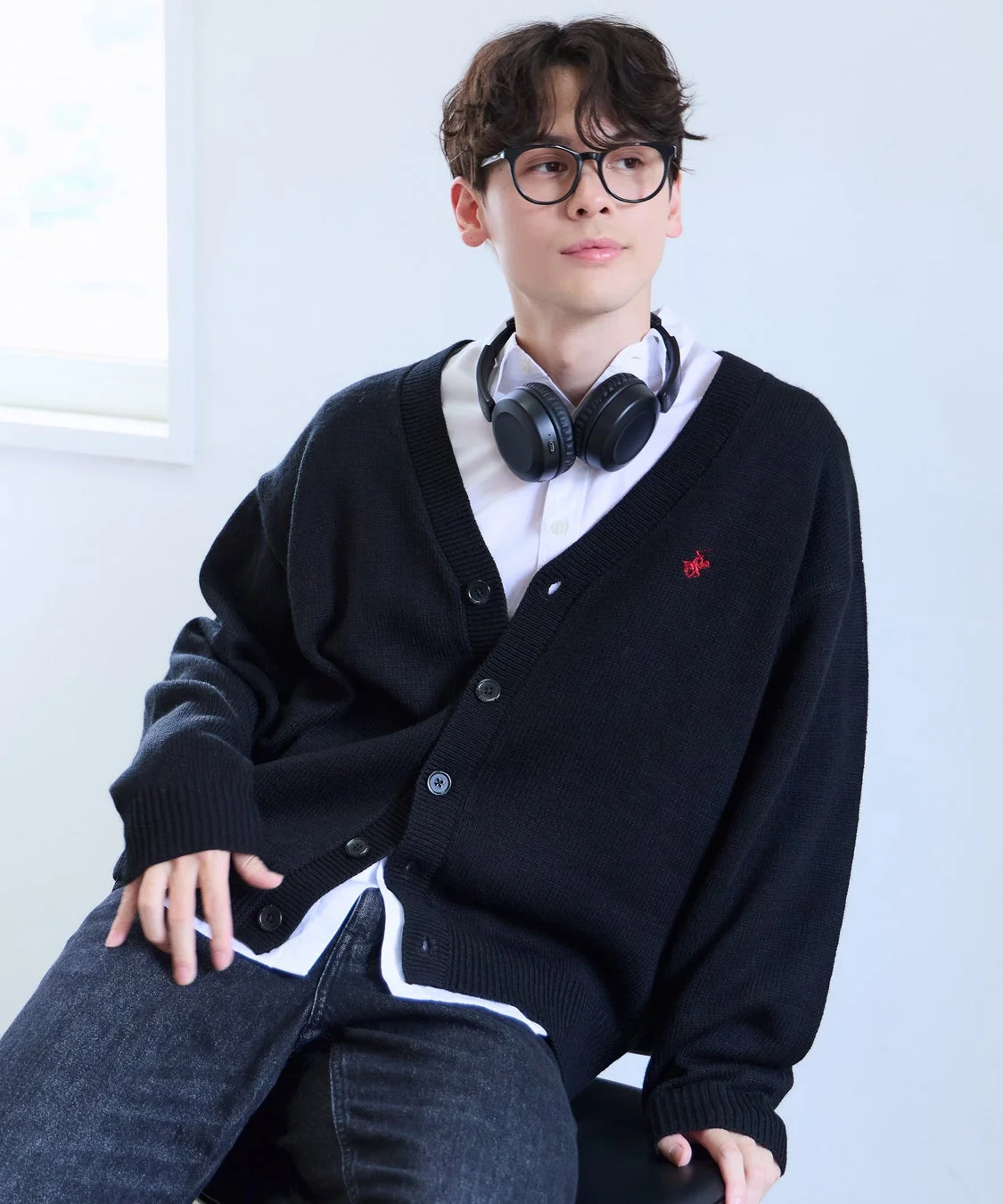 白いシャツと黒いカーディガンを着用し、眼鏡とヘッドホンを身につけた若い男性が、白い背景の部屋で座っている