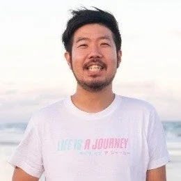 白いTシャツを着て笑顔で写るアジア人男性のポートレートです。Tシャツには「LIFE IS A JOURNEY」と日本語のメッセージが書かれています。背景はぼやけていますが、屋外の明るい環境であることがわかります。