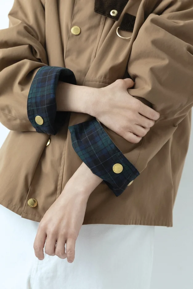茶色のジャケットを着た人物のクローズアップ画像。袖口から見えるタータンチェック柄と金色のボタンが特徴的で、カジュアルながらも上品な秋冬のファッションスタイルを表現しています。