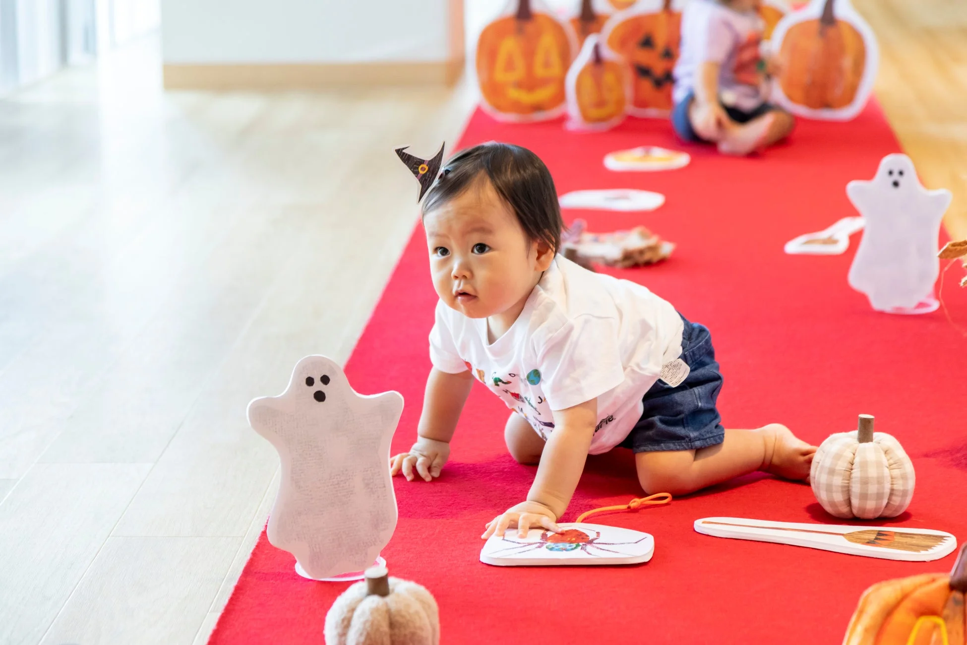 ハロウィンの飾り付けがされた赤いカーペットの上を、魔女の帽子をかぶった赤ちゃんが這い這いしています。