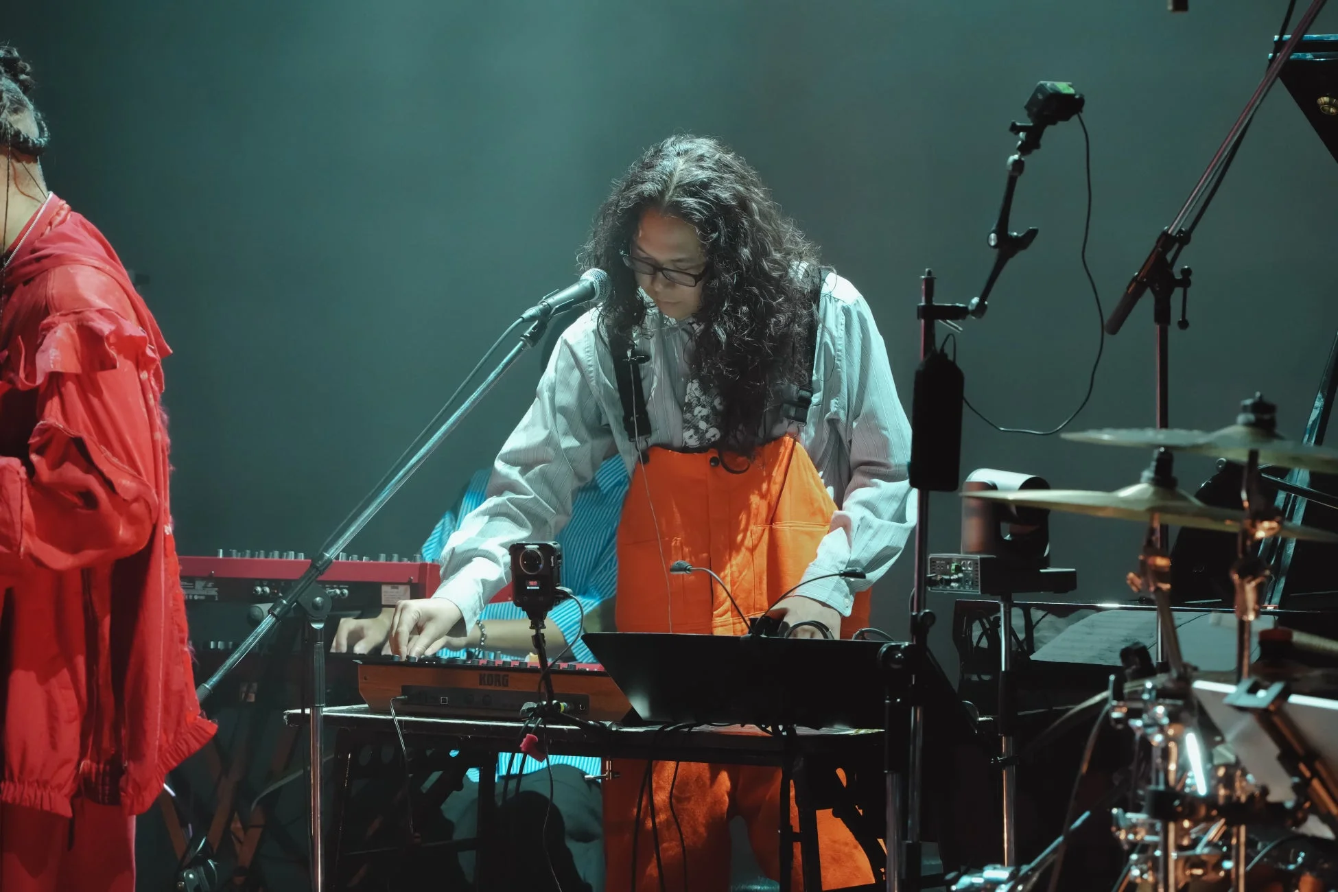 長髪と眼鏡のミュージシャンが、ライブステージでKORGのキーボードを演奏している様子。