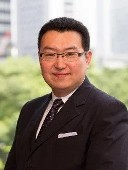 眼鏡をかけたアジア系の男性が、紺色のスーツとドット柄のネクタイを着用し、白いポケットチーフを差して笑顔でカメラを見つめているポートレート写真です。背景はぼかされており、ビジネスシーンを想起させます。
