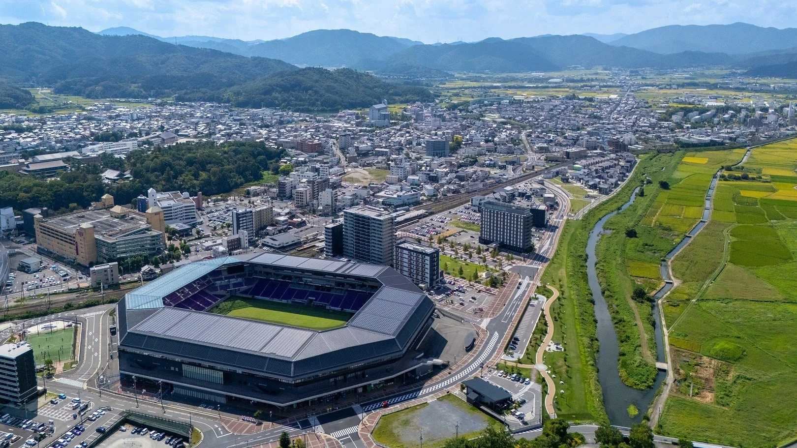 競技場を中心に、山々や田園風景が広がる日本の都市の空撮写真です。都市開発と自然が調和した景観が特徴的です。