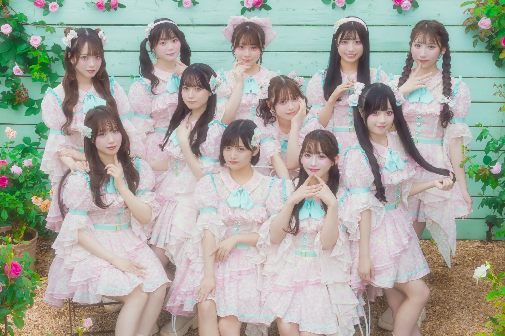アイドルグループと思われる複数の若い女性が、可愛らしいピンクと水色のフリルドレスを着て庭園でポーズをとっています。