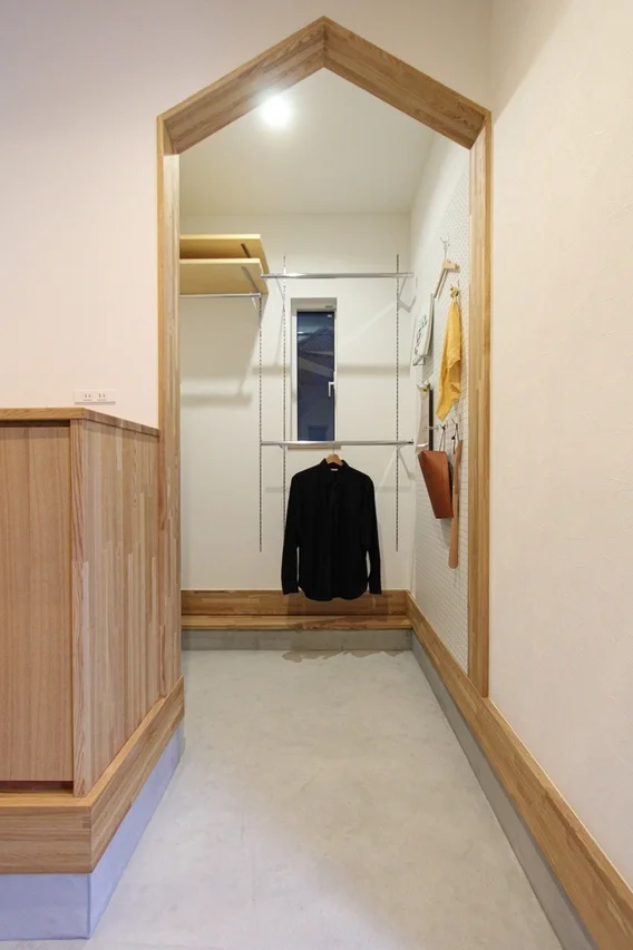 木製のアーチが特徴的な、モダンでミニマリストな玄関土間収納スペース。白い壁とコンクリートの床がシンプルさを際立たせ、木製家具、棚、ハンガーラックが機能的に配置されています。