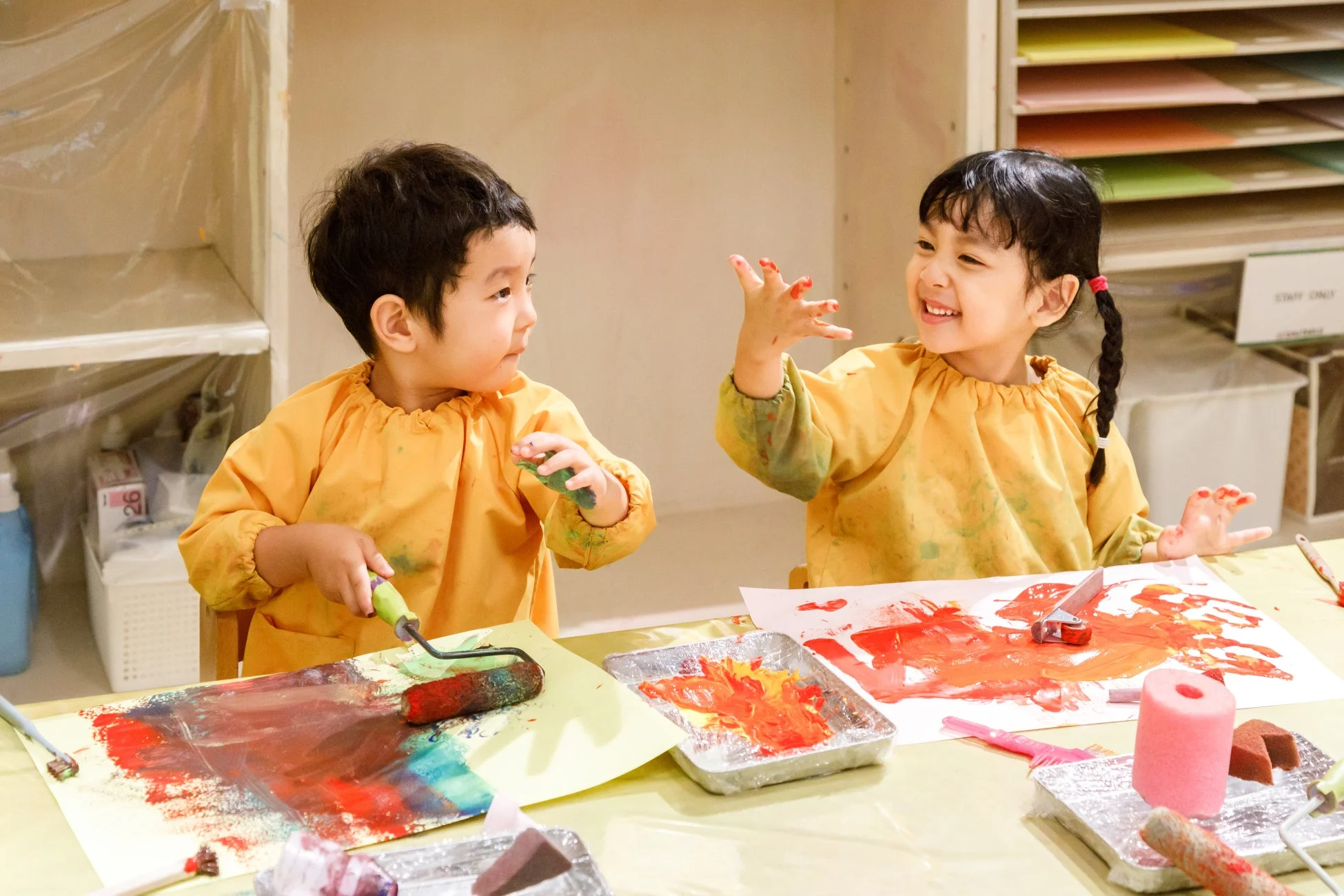 黄色いスモックを着た2人の幼児が、室内で絵の具を使って楽しそうにアート活動をしている。
