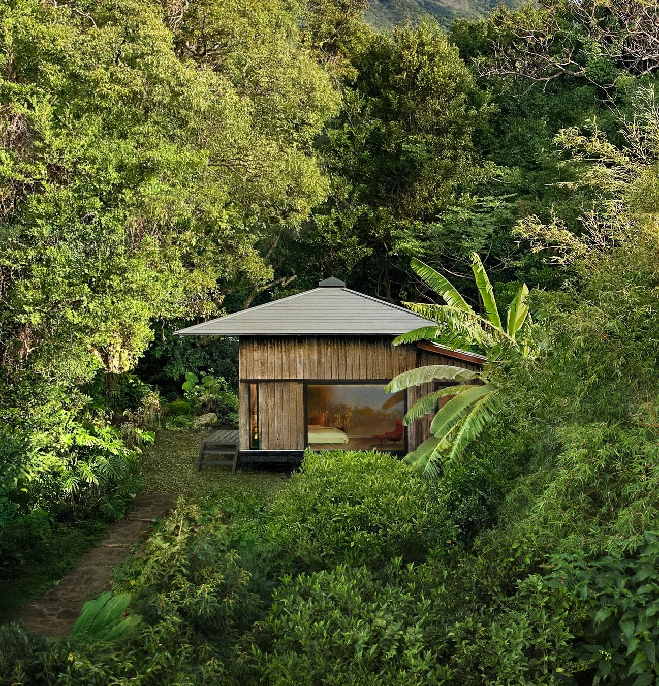 豊かな緑に囲まれた森の中に佇む木造の小屋の風景です。大きな窓からは室内の様子がうかがえ、静かで落ち着いた隠れ家のような雰囲気を醸し出しています。小道が小屋へと続いています。