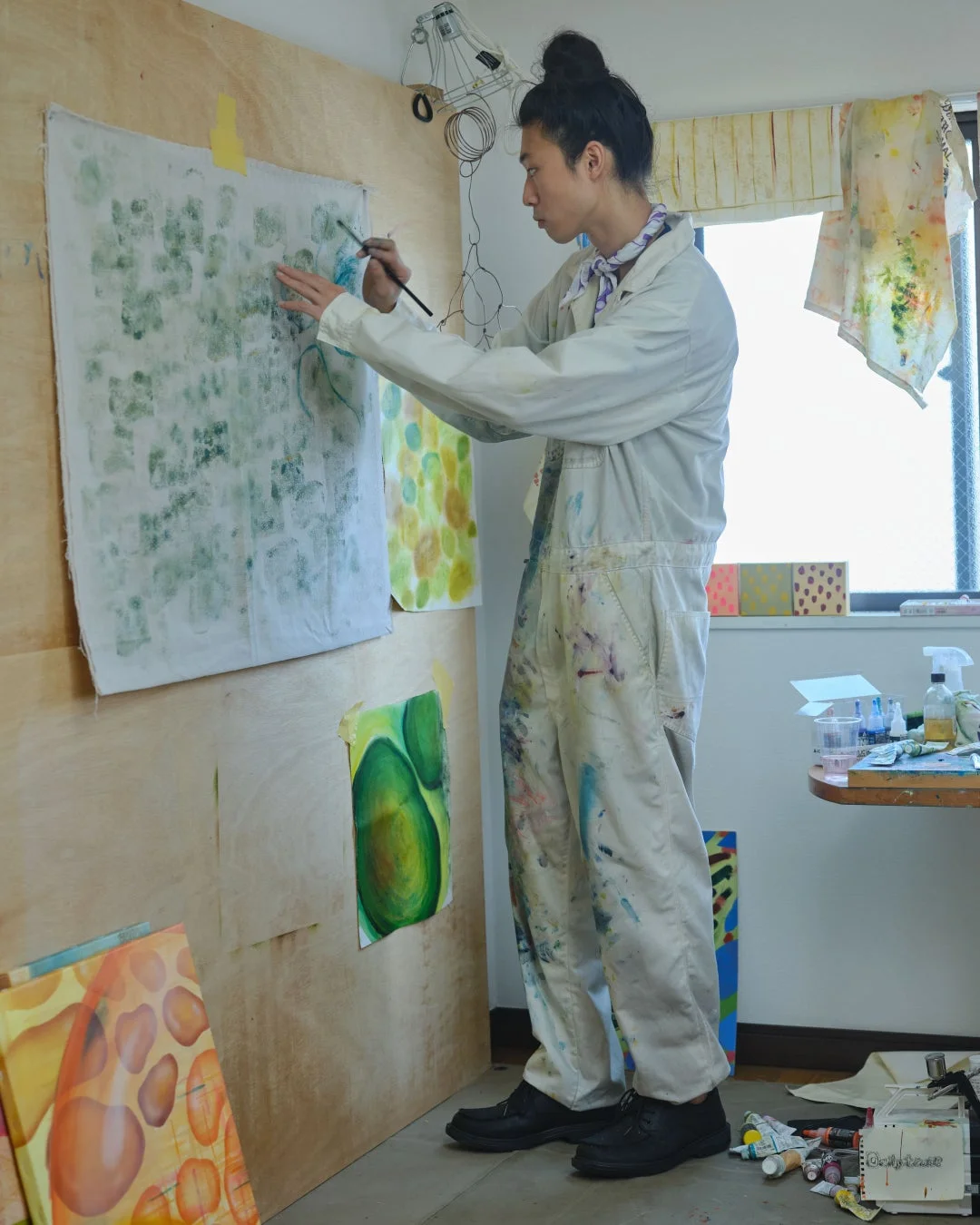 絵の具で汚れたオーバーオールを着た男性が、木製の壁に貼られたキャンバスに集中して絵を描いています。アートスタジオのような空間で、複数の作品が飾られています。