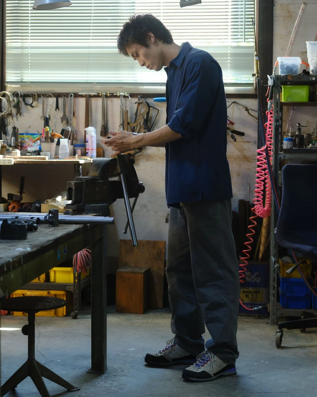 作業場でベンチバイスを使って何らかの作業に集中する若い男性の姿。壁には多くの工具が掛けられ、周囲には様々な道具が置かれており、ものづくりの環境が感じられる。