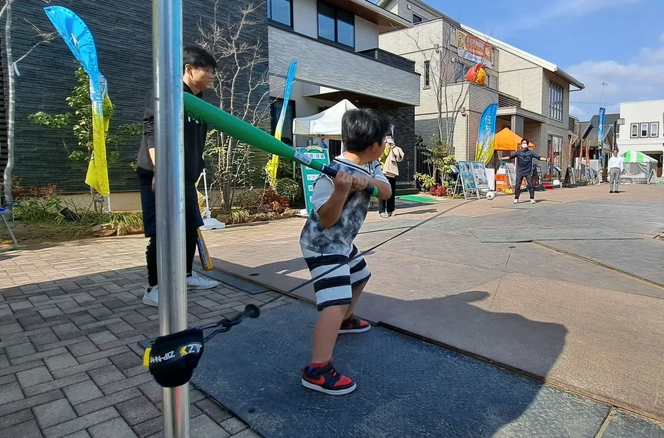 住宅街で行われている屋外イベントで、一人の子供が大人用の大きなバットを使って野球のバッティングゲームを楽しんでいます