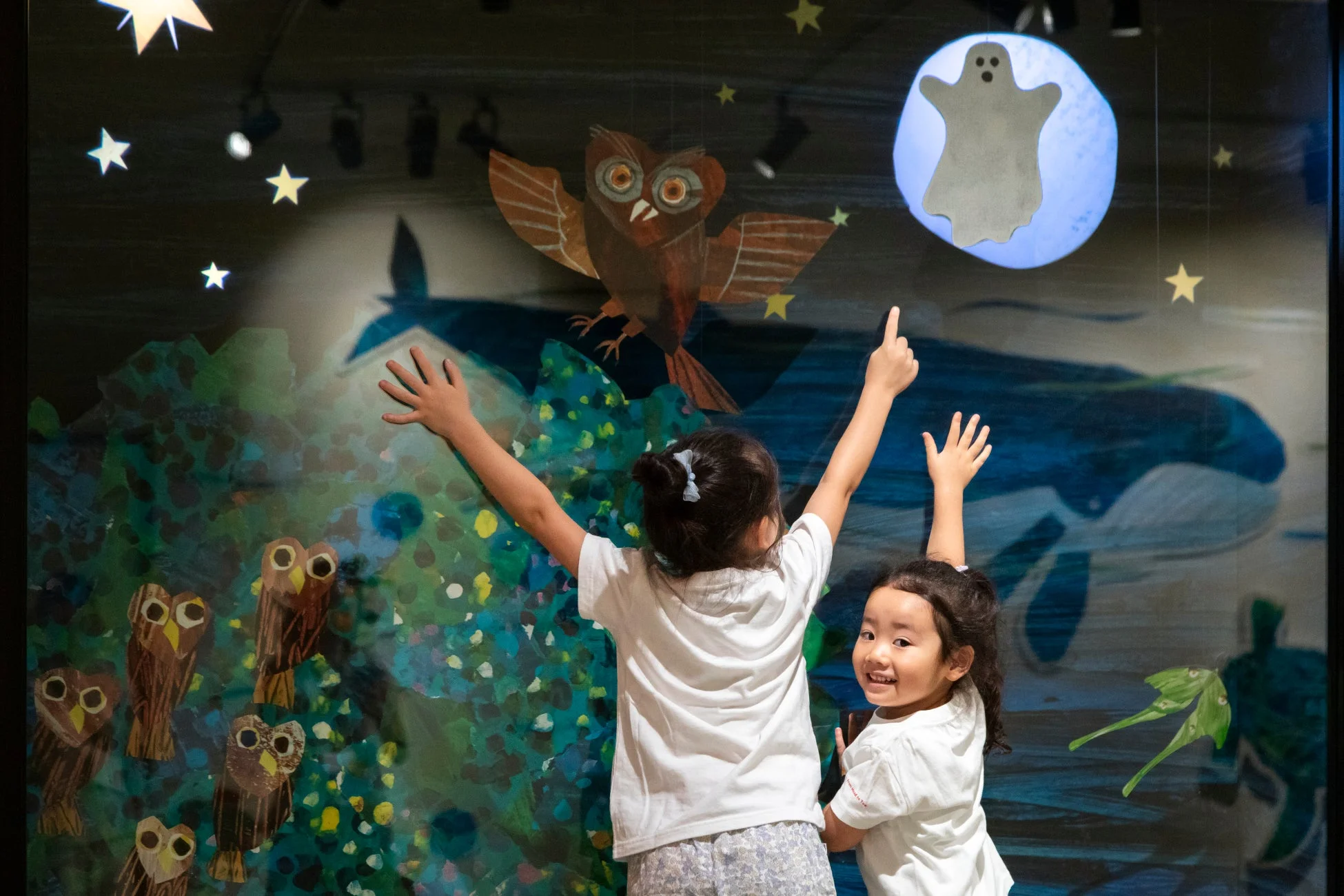 2人の幼い女の子が、フクロウやクジラ、星、幽霊が描かれた幻想的な夜空のディスプレイの前で、手を上げて楽しんでいる。
