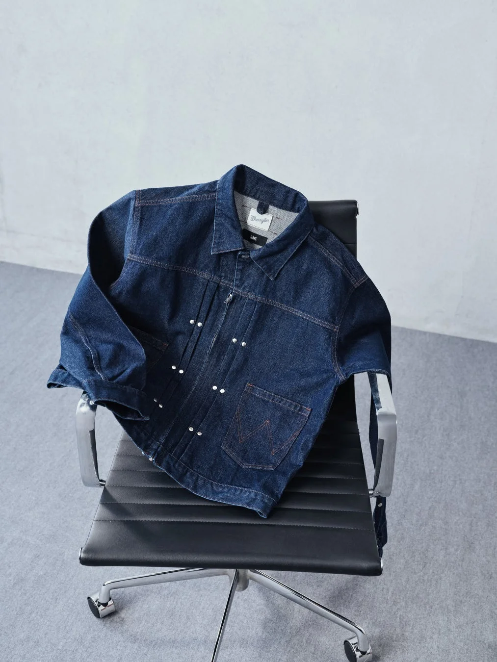 ダークブルーのデニムジャケットが黒いオフィスチェアにかけられています。ジャケットの襟元には「Wrangler」と「HARE」のブランドタグが見えます。シンプルながらもスタイリッシュなファッションアイテムの写真です。