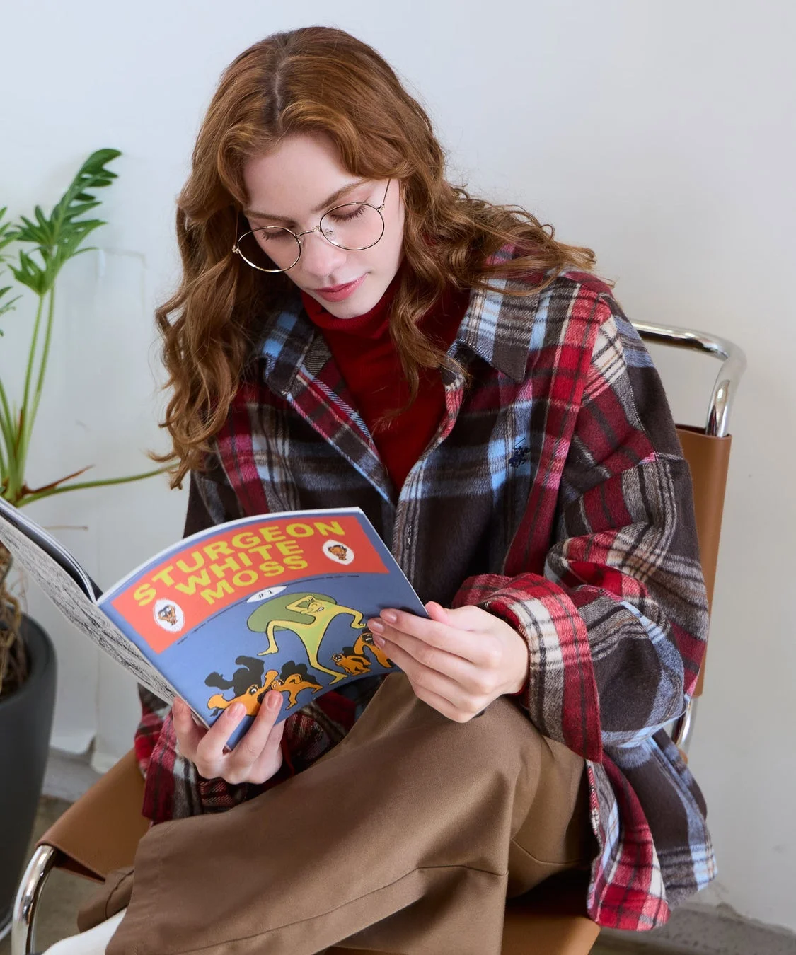 メガネをかけた女性がチェック柄のシャツを着て、椅子に座って本を読んでいる