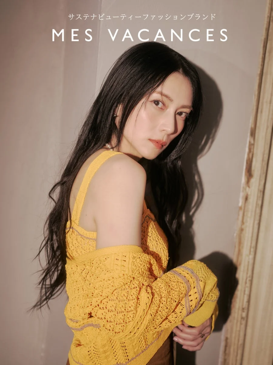 黒髪の女性が黄色のニットのトップスとカーディガンを着用し、カメラを見つめているポートレート写真。
