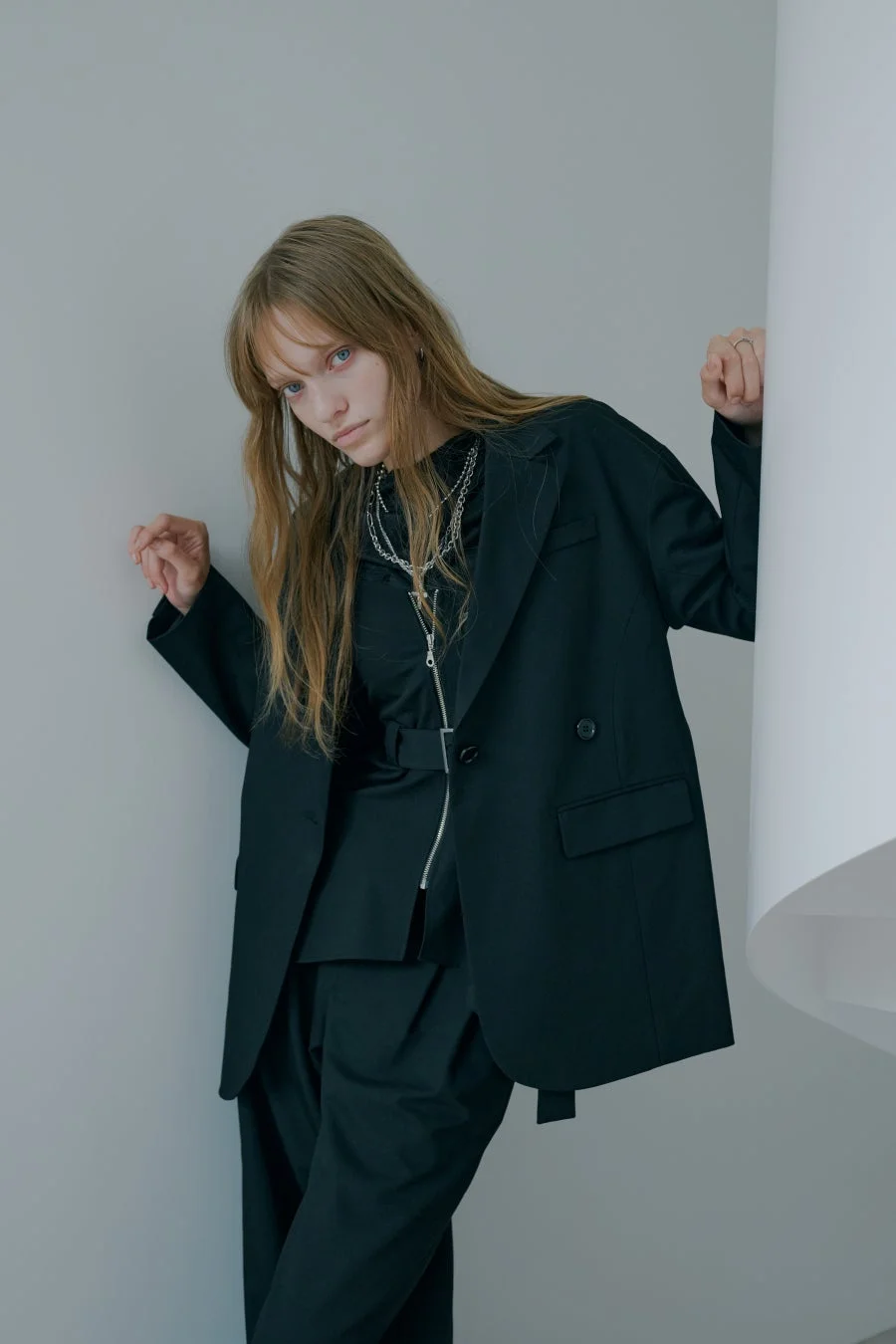黒いオーバーサイズのスーツを着た女性が、壁に寄りかかってポーズをとっています
