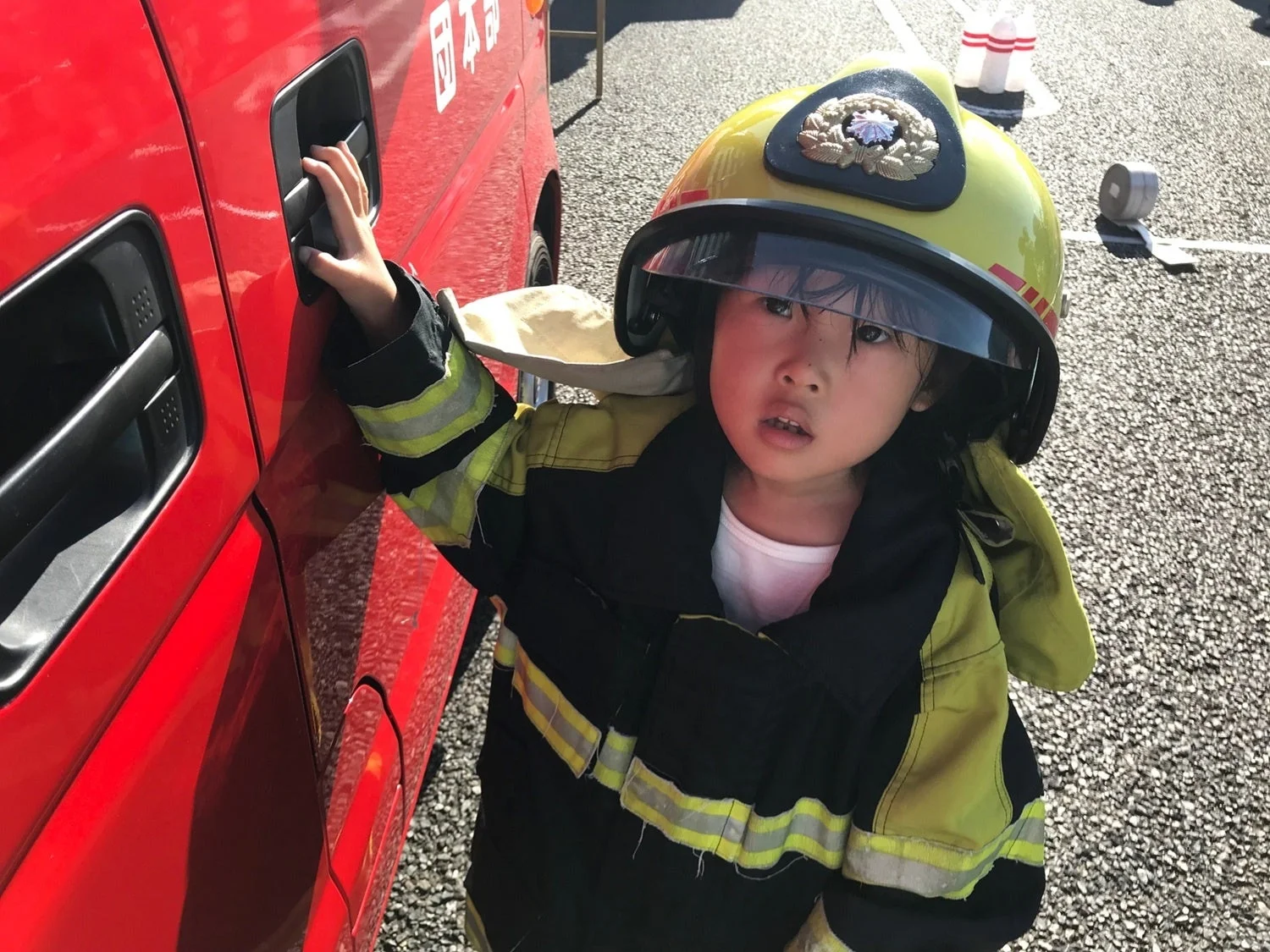 消防士のヘルメットと制服を着用した幼い子供が、赤い車両のドアハンドルに手を伸ばしている
