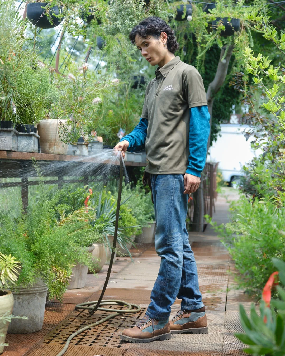緑豊かな温室で、男性がホースを使って植物に水やりをしている様子。リラックスした雰囲気の中で、手入れが行き届いた植物たちが並んでいます。