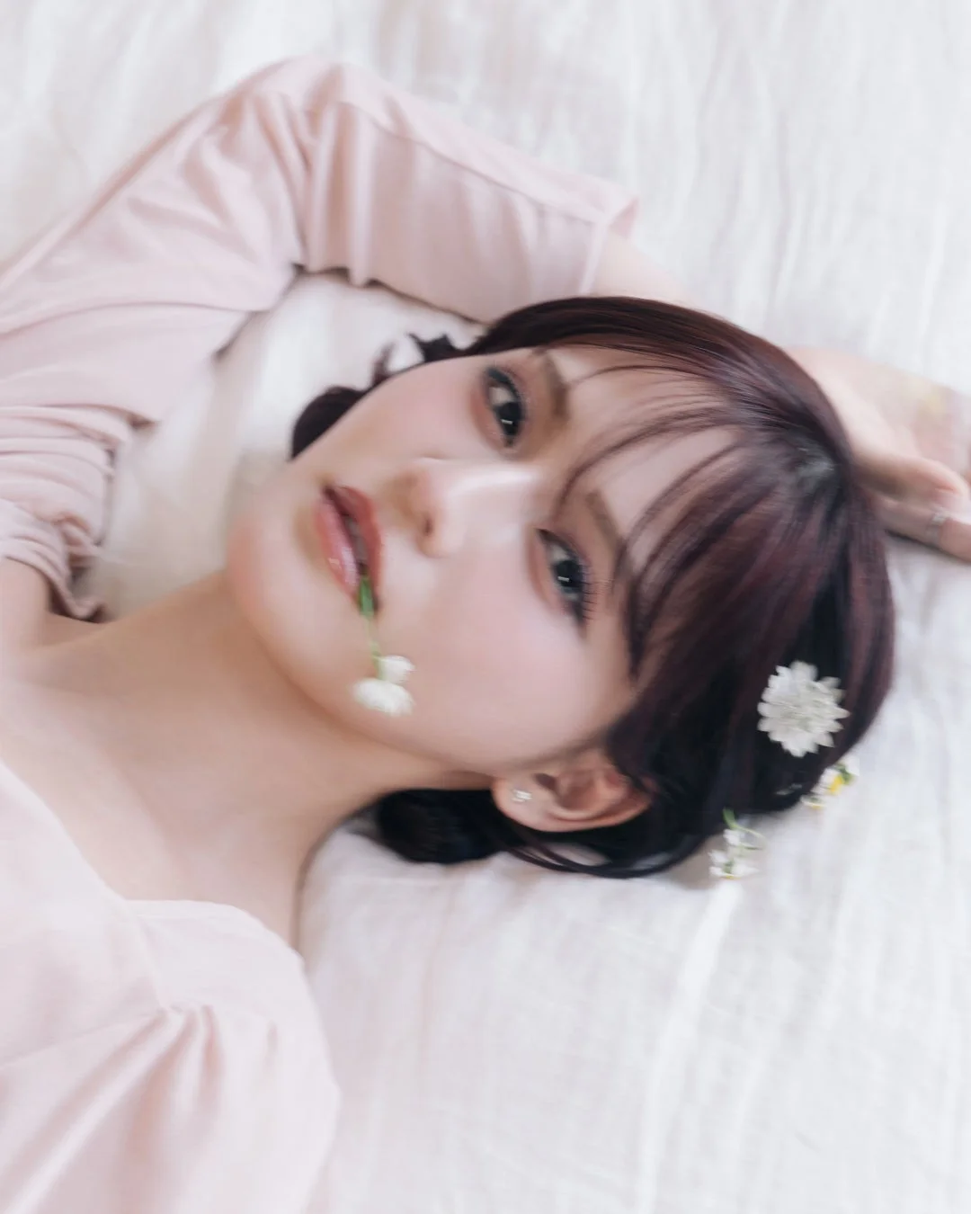 ピンク色の服を着た女性が白いシーツに横たわり、髪や口元に小さな白い花を飾っているポートレートです。