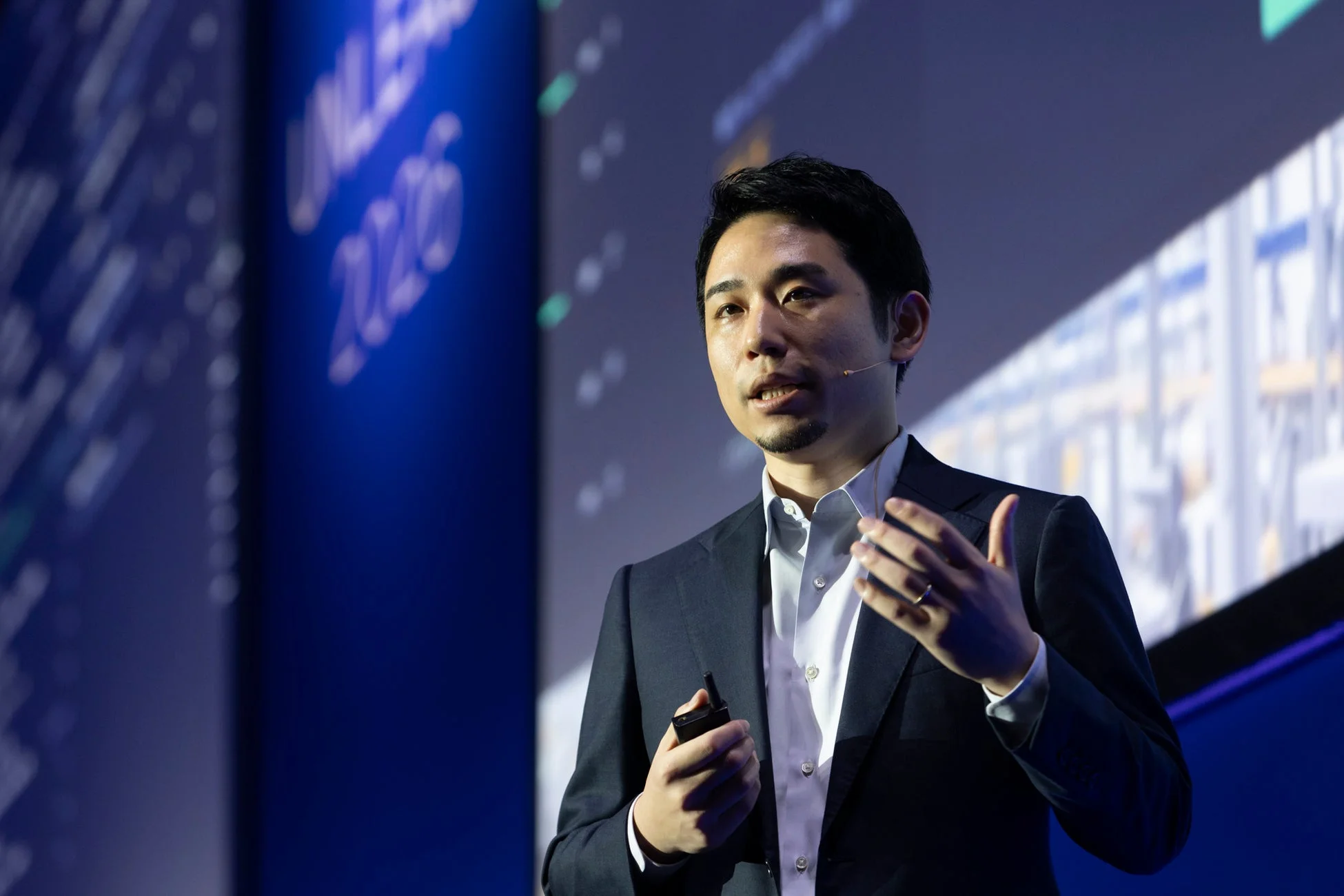スーツを着た男性がステージ上でプレゼンテーションを行っています。背景のスクリーンには「UNLEASH 2020」の文字が見え、イベントでの講演風景を捉えた一枚です。