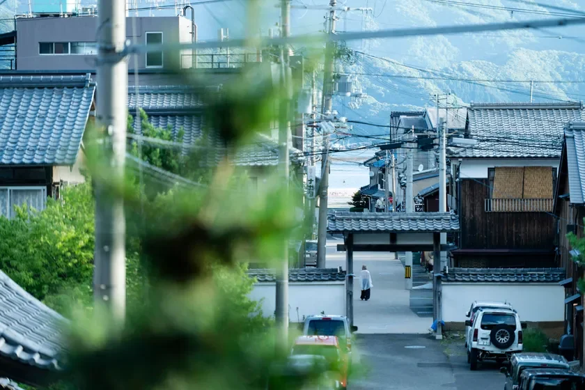 瓦屋根の家々が並ぶ日本の古い街並み。電柱や電線が張り巡らされ、奥には海と山が見えます。門をくぐる人物の姿が印象的な、どこか懐かしさを感じる風景です。