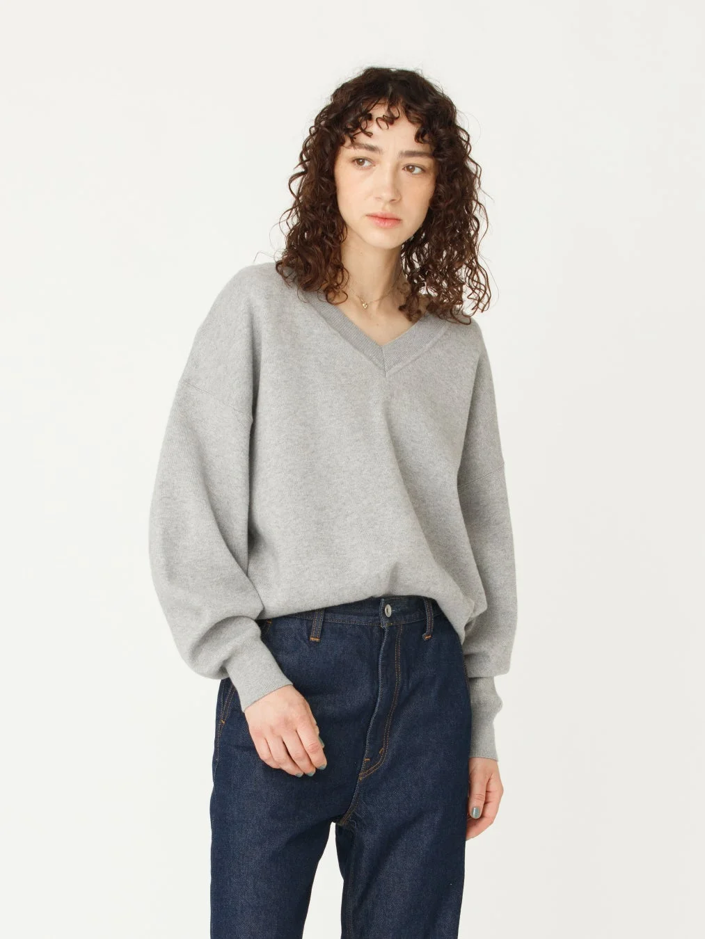 白い背景の前で、カーリーヘアの女性がグレーのVネックニットとデニムパンツを着用しています。落ち着いた雰囲気のカジュアルな着こなしです。