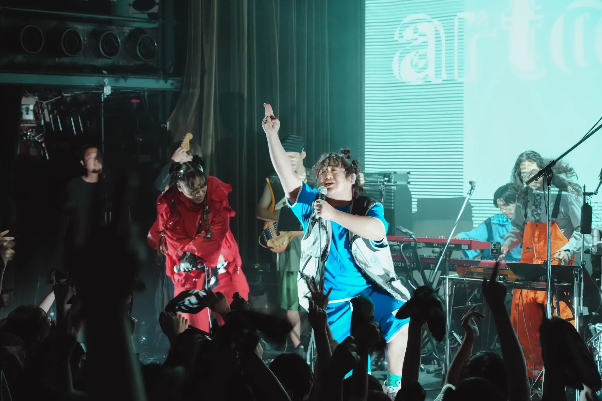 ライブステージでバンドが熱演しており、中央のボーカルがマイクを手に観客を煽っている。