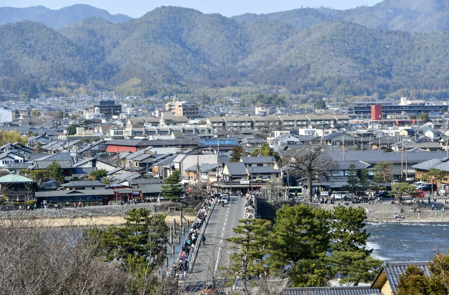 京都の嵐山にある渡月橋と桂川、そしてその周辺の街並みを遠景で捉えた写真です。多くの観光客が橋を行き交い、背景には連なる山々が広がっています。
