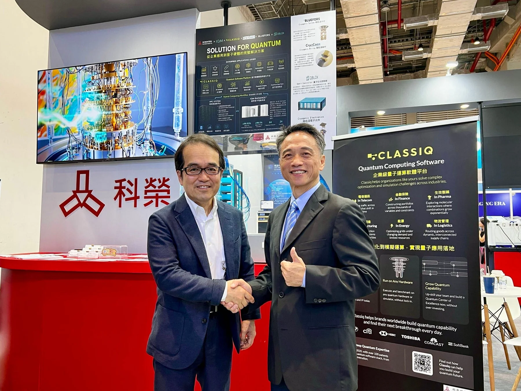 量子コンピューティングの展示ブースで、二人のビジネスマンが握手している様子です。Classiqの量子計算ソフトウェアや、様々な企業による量子ソリューションが紹介されており、技術提携やビジネス協業の場面を示しています。