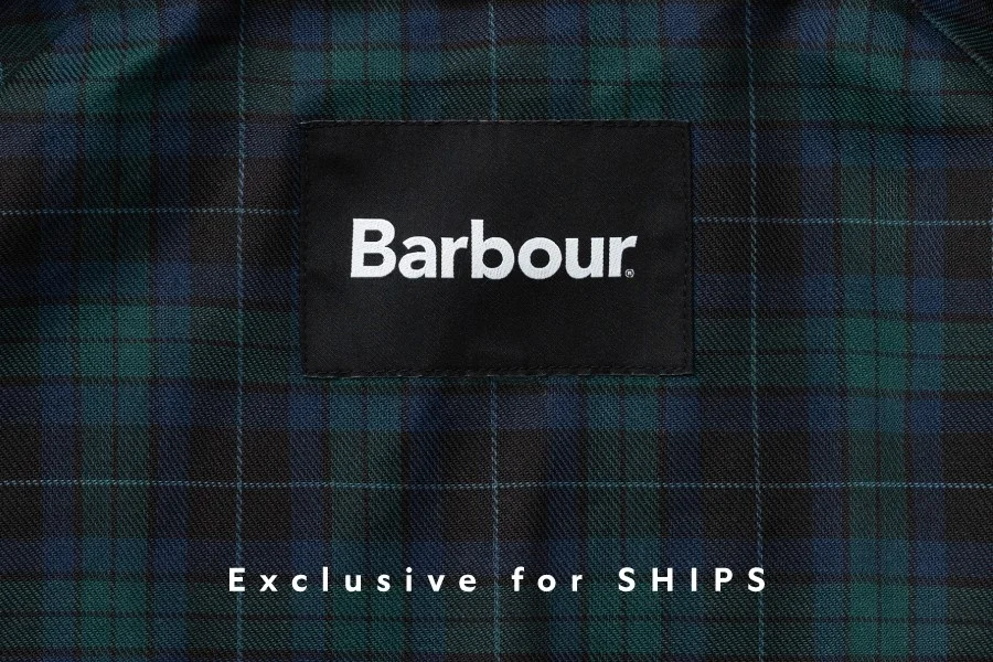 Barbourブランドのロゴが入った黒いラベルが、緑と青のタータンチェック柄の生地に縫い付けられています。下部には「Exclusive for SHIPS」と書かれており、SHIPSとの限定コラボ商品であることを示しています。