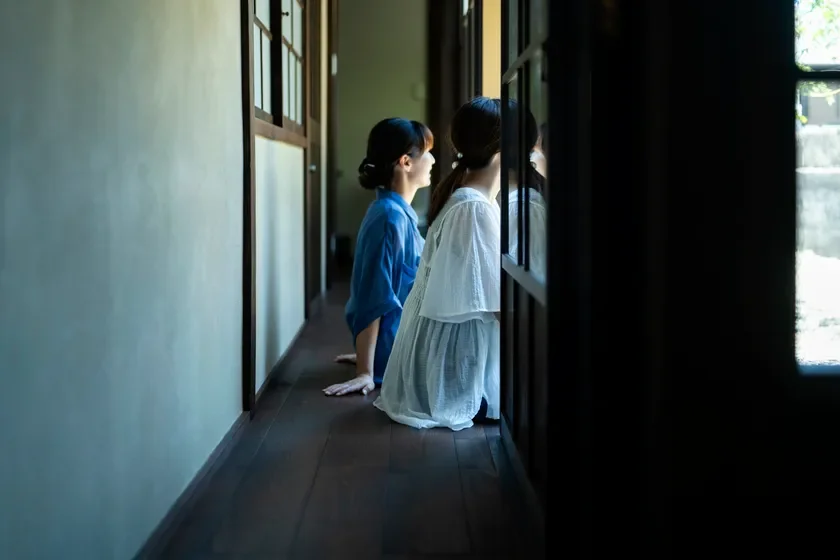 廊下の床に座り、窓の外を眺める二人の女性の横顔を捉えた画像です。静かで落ち着いた雰囲気の中で、二人が何かに思いを馳せているかのような情景が描かれています。