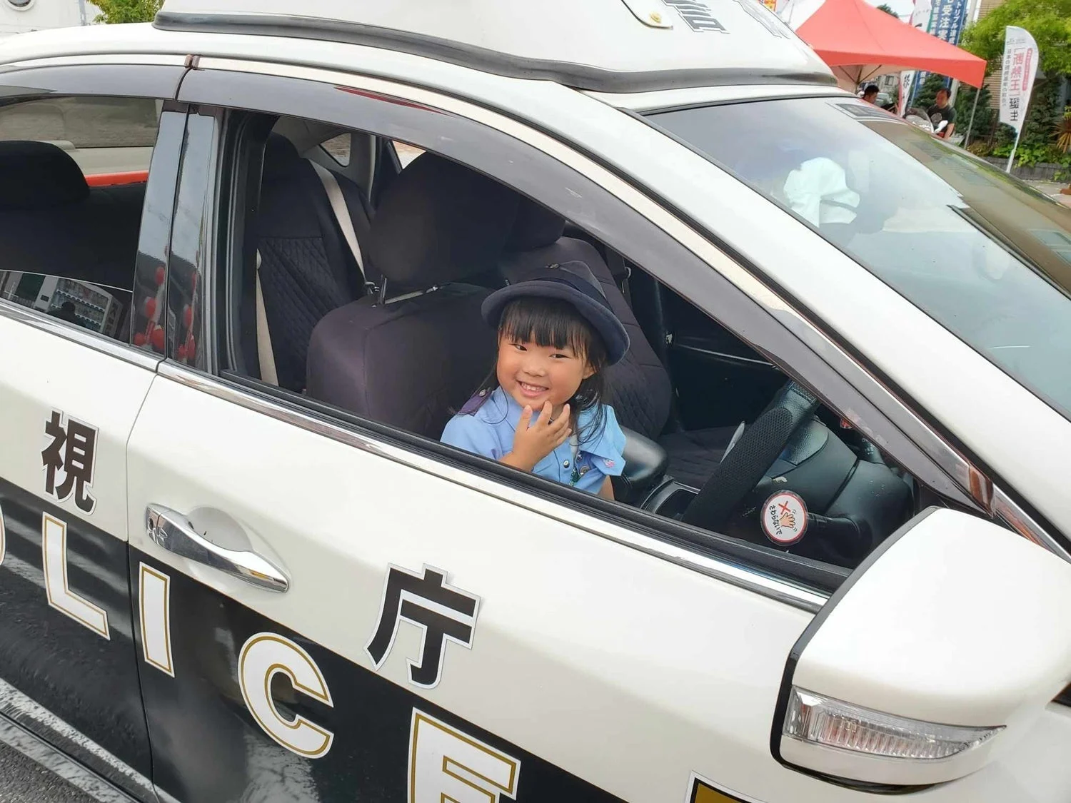 警察官の制服を着た小さな女の子が、パトカーの運転席に座って笑顔を見せる